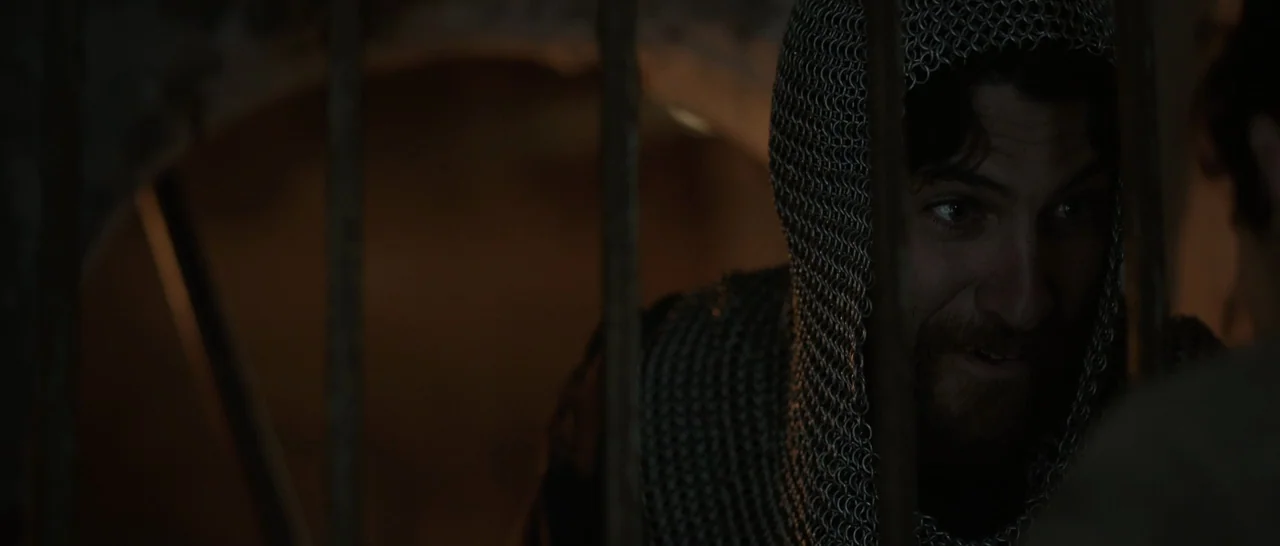The Little Hours (2017) movie still: closeup — A dimly lit dungeon space features vertical iron bars separating the subject. A man wearing a…