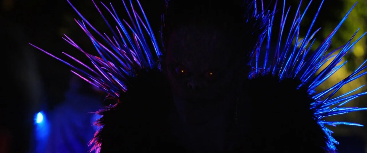 Death Note (2017) movie still: closeup — A dark night street setting contains a creature with glowing yellow eyes and long spikes. The creature…