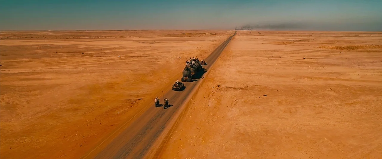 Mad Max: Fury Road (2015) movie still: wide — A vast, arid desert stretches to the horizon with a single straight asphalt road. A convoy of heavily…