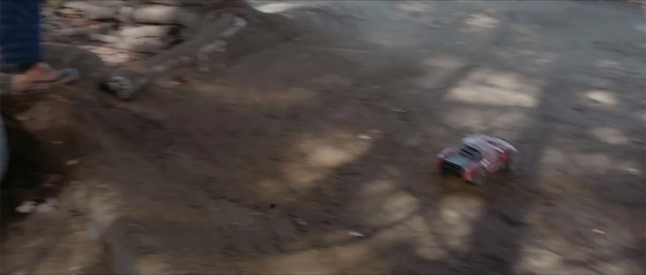 Bludgeon (2018) movie still: wide — A muddy outdoor trail is shaded by trees with light dappled across the ground. A small red remote-controlled car…