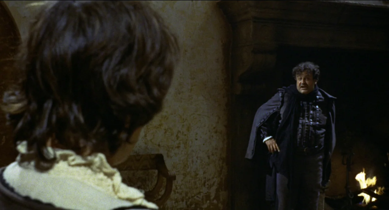Beatrice Cenci (1969) movie still: medium — A stone chamber features textured walls and a large, recessed hearth containing an active fire. An older…