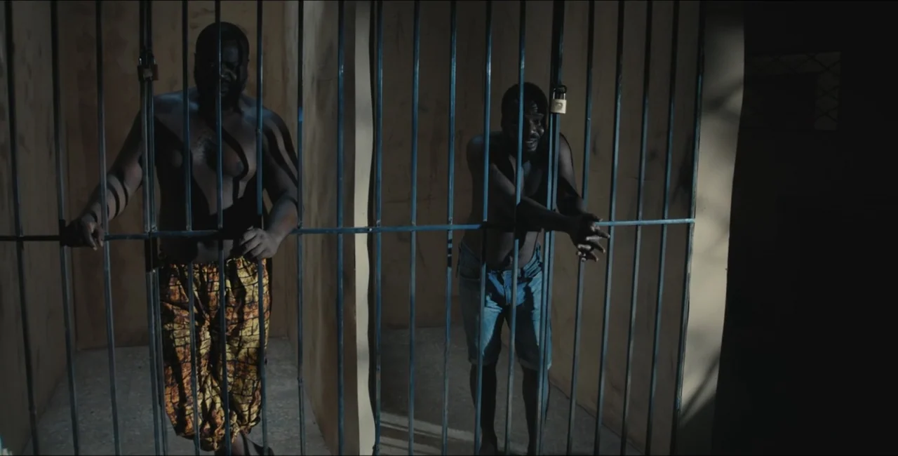 Project Assault (2025) movie still: fullbody — A small room with beige concrete walls enclosed by thick vertical metal bars. Two shirtless men stand…