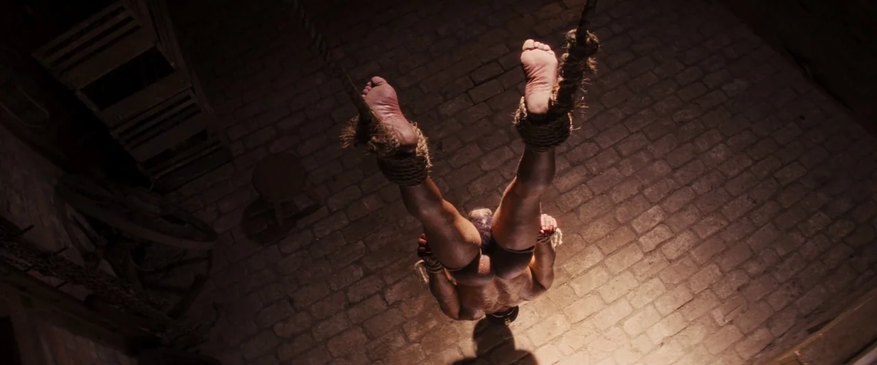 Django Unchained (2012) movie still: medium — A dark subterranean space with stone walls and a floor made of irregular bricks. A muscular man hangs…