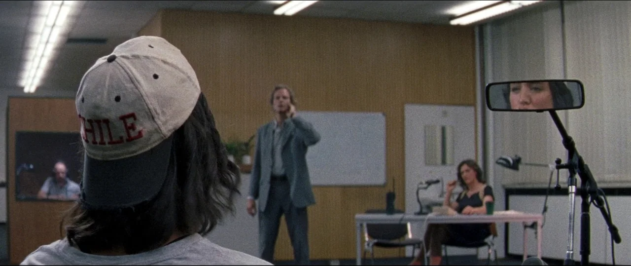 Luz (2018) movie still: medium — A workspace with wood-paneled walls and fluorescent lights contains several people at desks. A man in the…