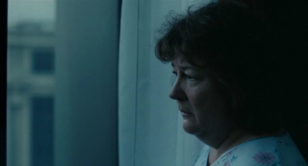 Paris, Je T’Aime (2006) movie still: closeup — A dimly lit room with a sheer curtain partially obscuring an exterior view. A woman stands by the…