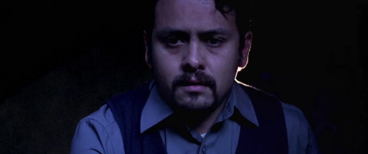 Here Comes The Devil (2012) movie still: closeup — A man stands in an underexposed, dark environment with no visible surroundings. He looks directly…