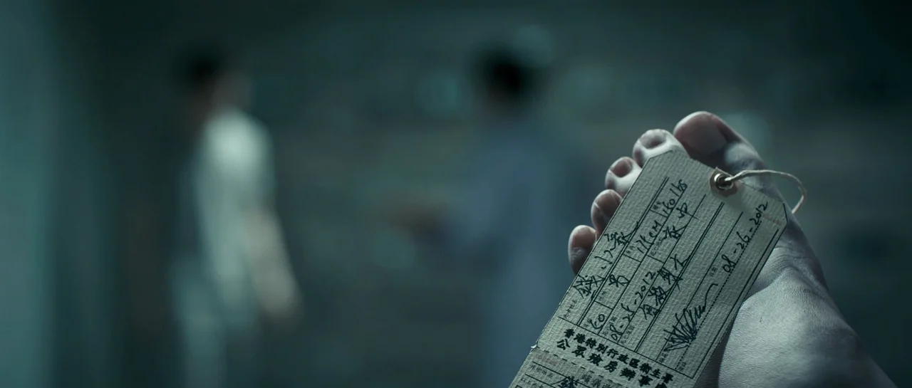 Rigor Mortis (2013) movie still: closeup — A close up of a paper medical toe tag attached to a human foot. Two blurred figures in scrubs stand in…