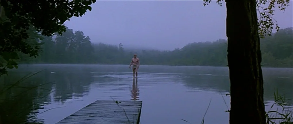 IP5 (1992) movie still: wide — A misty, calm lake is surrounded by dense trees on a foggy morning. An elderly man walks chest-deep through the…