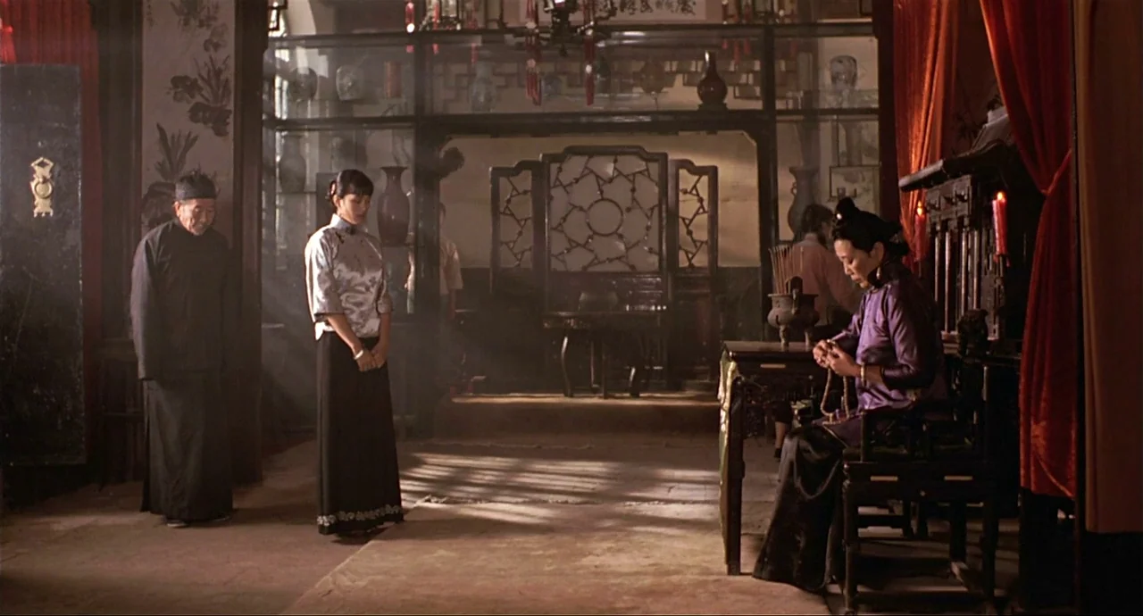 Raise The Red Lantern movie still: wide — A traditional room with carved wooden screens and ceramic vases on display shelves. Three people stand or…