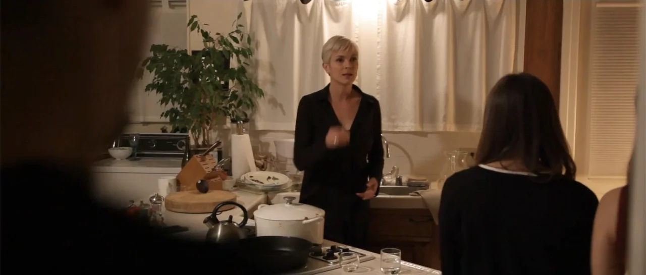 La coherencia del caos movie still: medium — A residential kitchen features white curtains, a countertop with cookware, and a potted plant. A woman…
