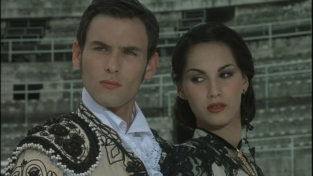 Romance (1999) movie still: medium — A stone stadium with tiers of seating forms the background behind two people. A man in a matador costume and a…