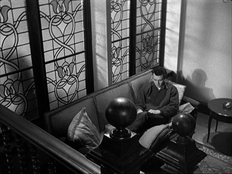 The Sleeping Tiger (1954) movie still: wide — A stairwell landing features a couch placed against tall leaded-glass windows with floral patterns. A…