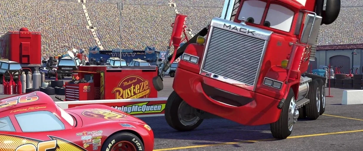 Cars (2006) movie still: medium — A sunny racetrack pit area features a red transport truck and a race car. The characters are positioned on asphalt…
