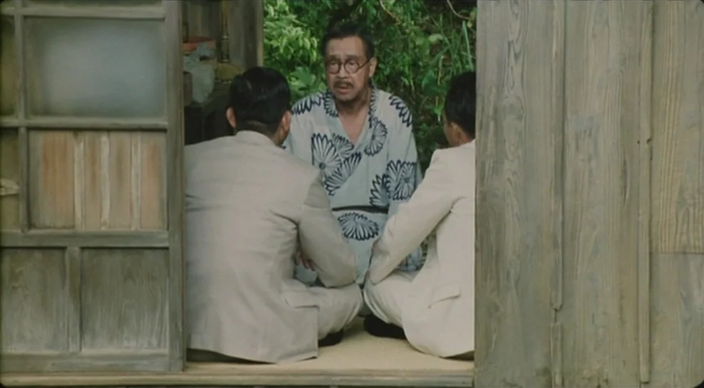 Madadayo (1993) movie still: medium — A traditional room with open sliding wooden doors overlooks a lush green outdoor area. Three men are seated on…