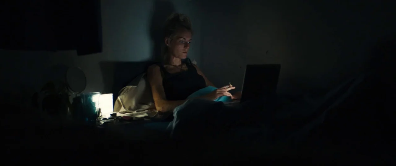 Silver Haze (2023) movie still: medium — A dark, dimly lit space where a person sits propped up in a bed. The individual types on a laptop computer…