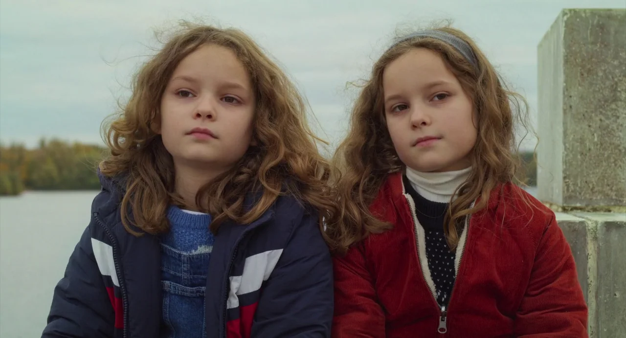 Petite Maman (2021) movie still: medium — Two young girls sit together outdoors with a body of water in the background. They both face forward with…