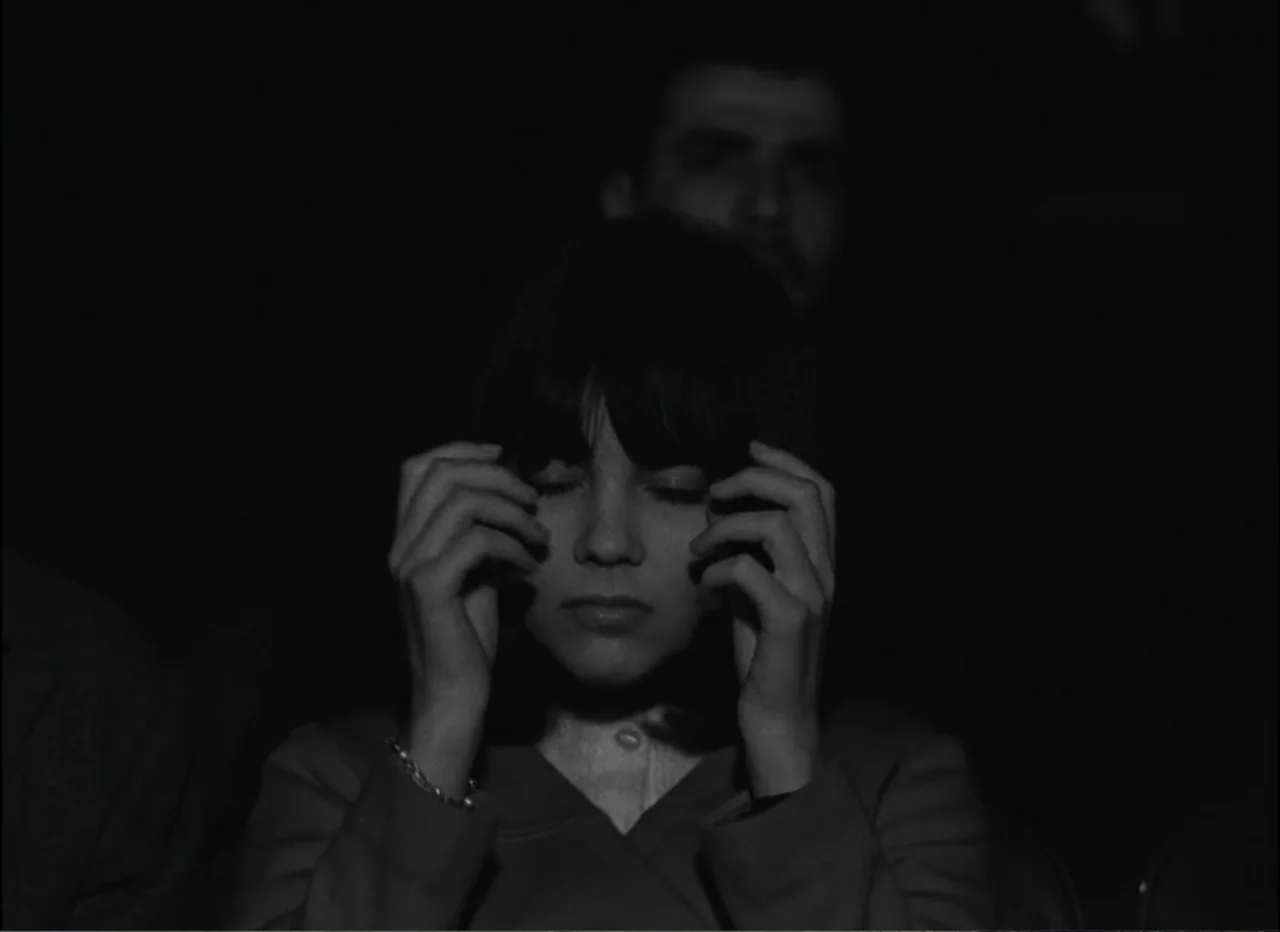 Masculin Féminin (1966) movie still: medium — A dark theater auditorium with a seated woman in the foreground and a man behind. The woman rests her…