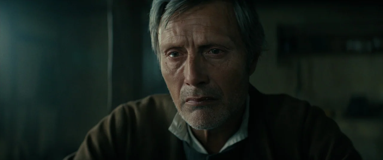 The Promised Land (2023) movie still: closeup — A dark, sparsely furnished interior with muted, cool-toned light filtering through a side window. A…