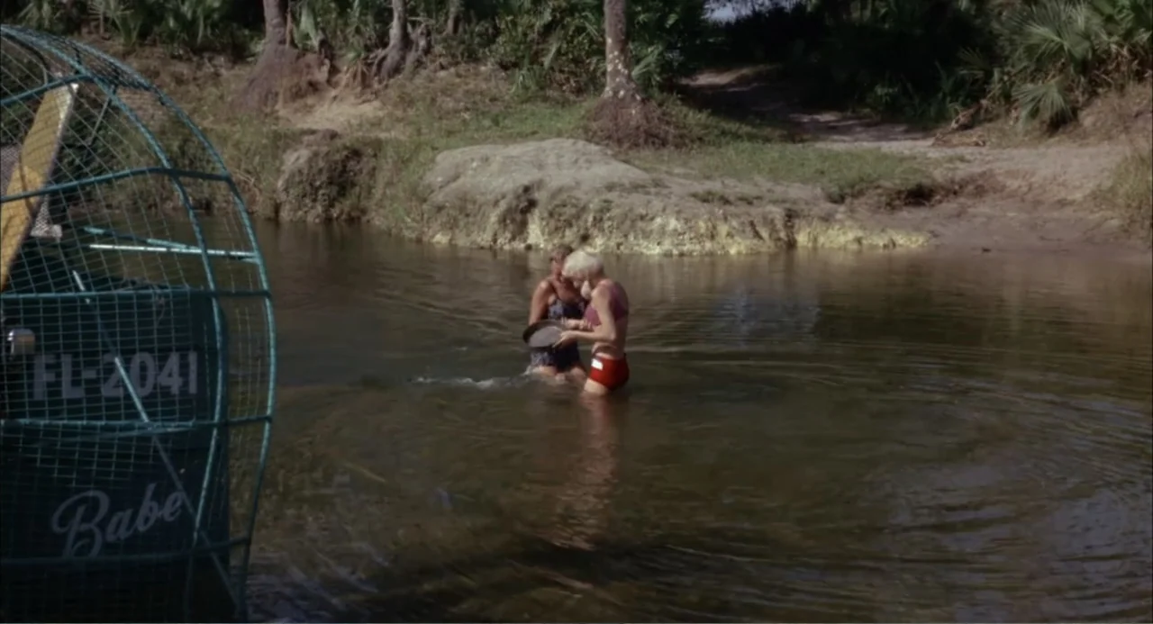 Cottonpickin' Chickenpickers (1967) movie still: medium — A murky river flows through a densely forested area with palm trees on banks. Two women in…