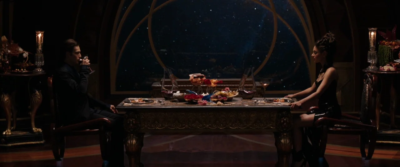 Jupiter Ascending (2015) movie still: wide — A dimly lit room features a formal dining table with a celestial background view. A man and a woman sit…