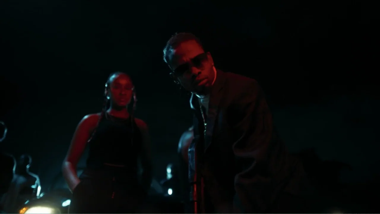 Gangs of Lagos (2023) movie still: medium — A dark outdoor urban space features a parked vehicle with its headlights casting red light. A man in a…