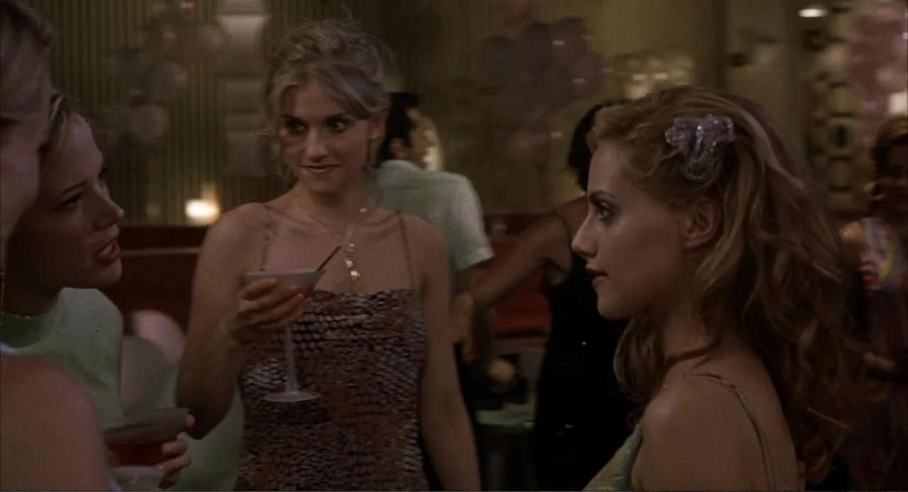 Uptown Girls (2003) movie still: medium — A dimly lit lounge setting features clusters of people gathered for a social event. Three women stand in a…