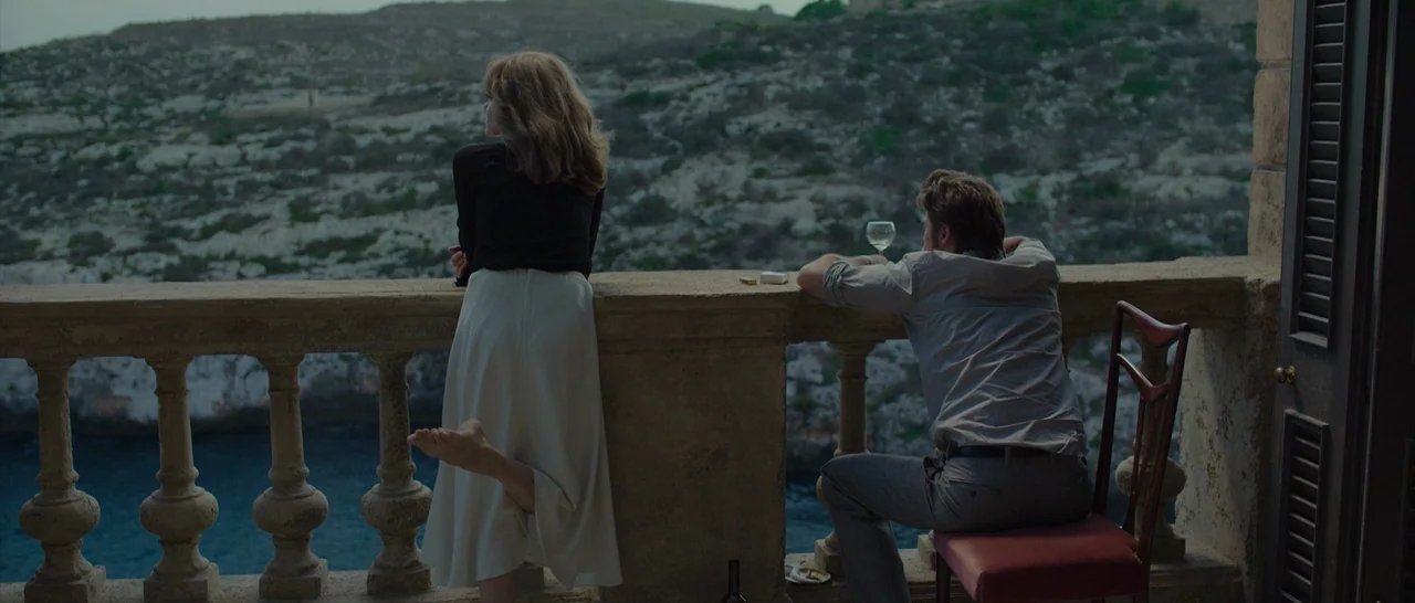 By The Sea (2015) movie still: wide — A stone balcony with ornate balusters overlooks a rugged cliffside and calm water. A woman leans against the…