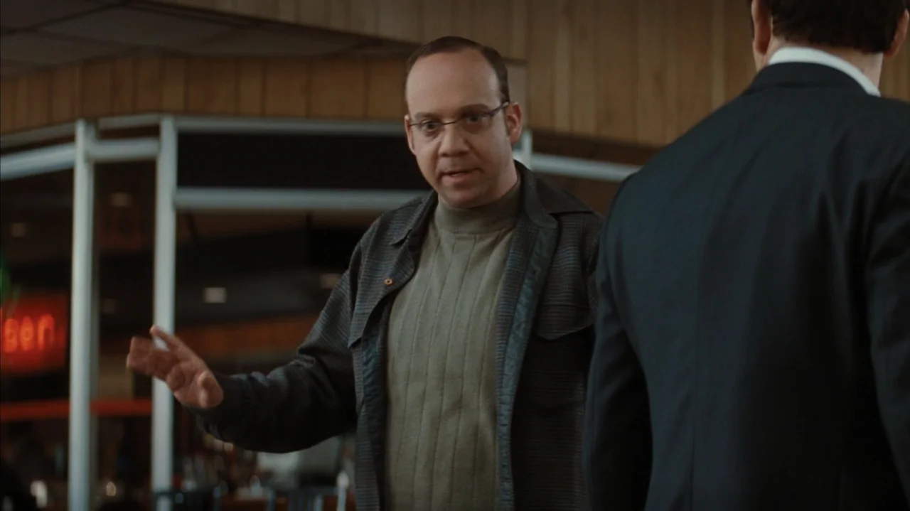 Duplicity (2009) movie still: medium — A diner booth interior features wooden wall paneling and blurred storefront windows. A man gestures while…