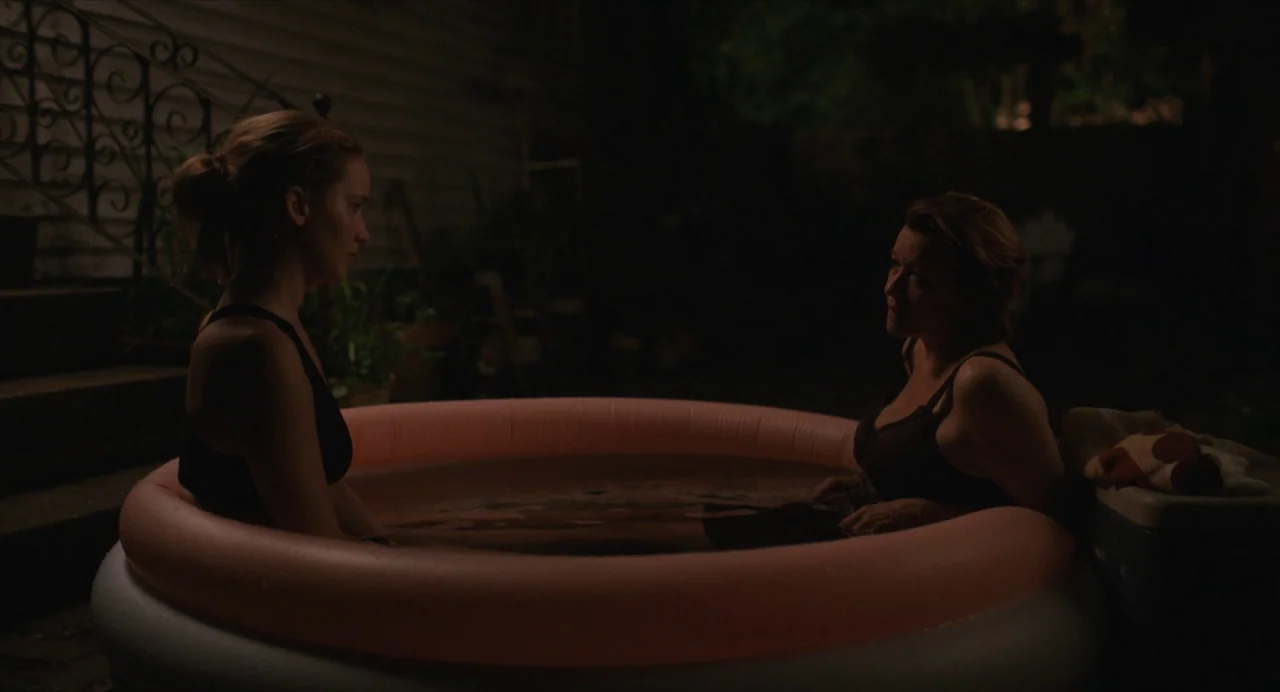 Causeway movie still: medium — An inflatable pink circular pool sits on a dark patio at night. Two women sit across from each other in the water…