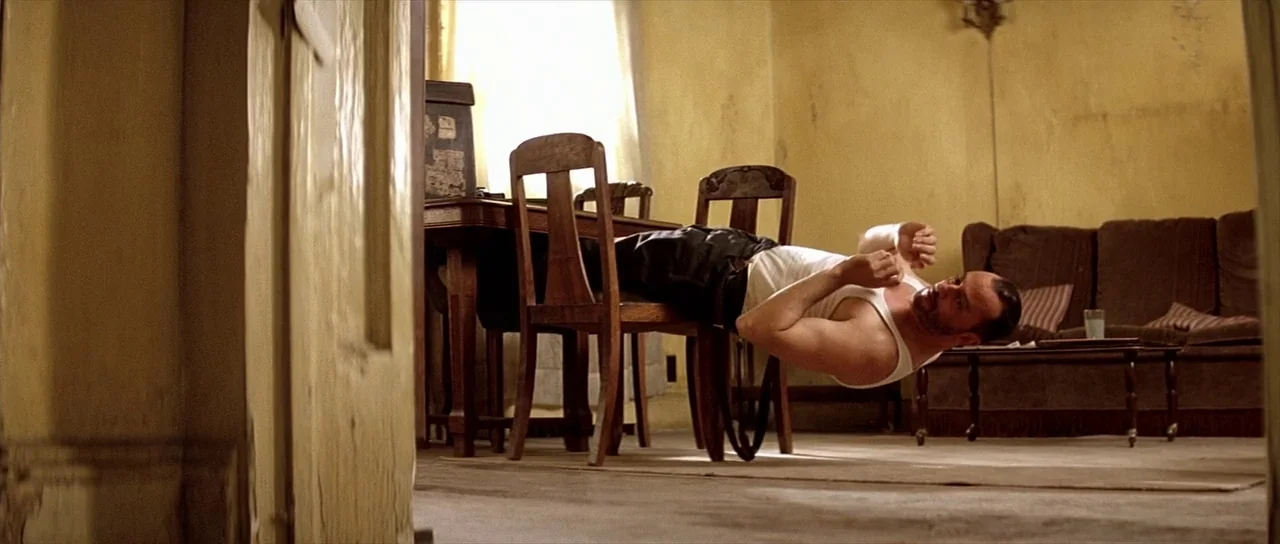 Léon: The Professional movie still: wide — A sparsely furnished room features weathered yellow walls and worn wooden furniture. A man holds a rigid…