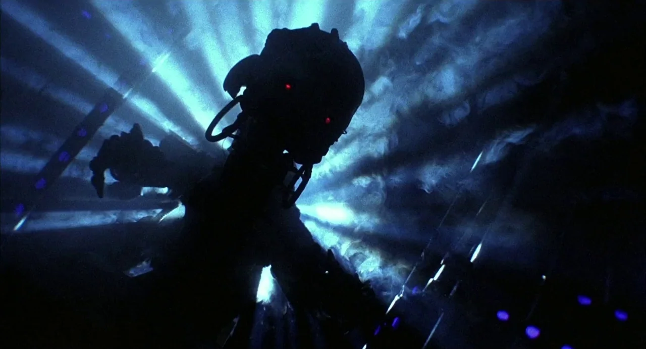 Hardware (1990) movie still: medium — A dark figure in a helmet stands in a smoke-filled, dimly lit environment. Strong blue light rays project from…