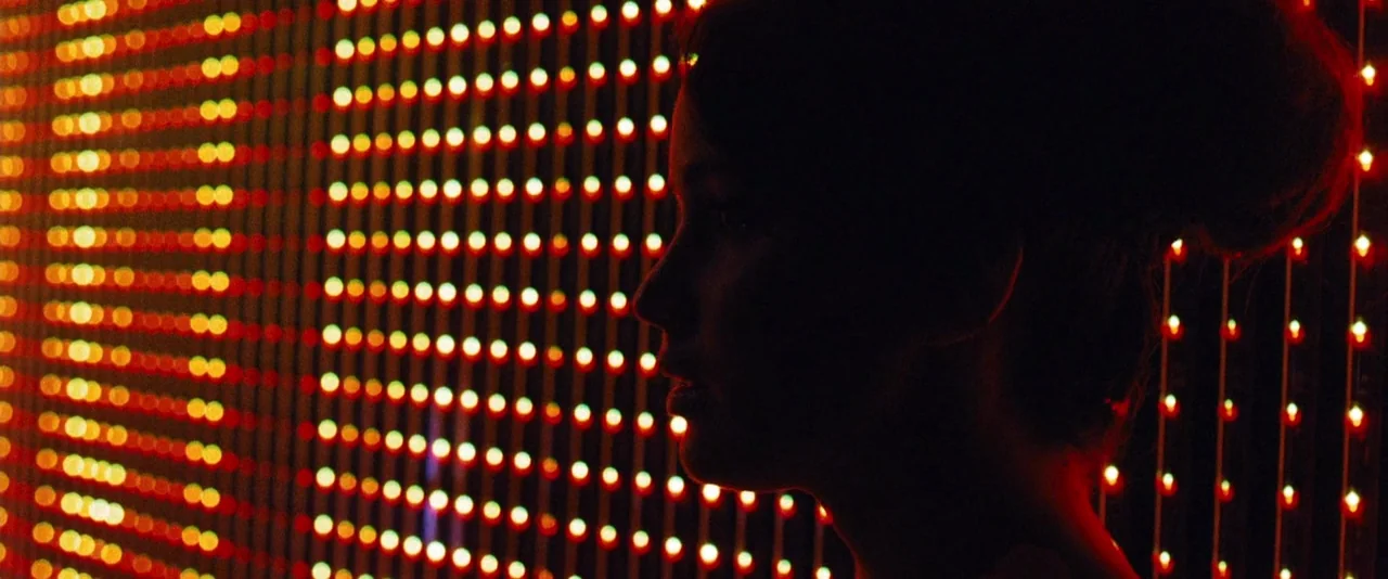 The Hunger Games (2012) movie still: closeup — A woman stands before a wall of vertical light strips in a dark room. The lights create a grid of…