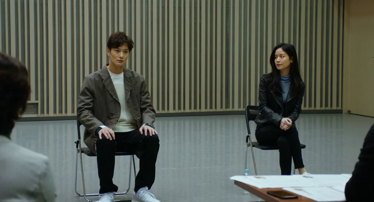 Drive My Car (2021) movie still: fullbody — A spacious room features vertical wooden slats on the back wall and gray floors. Two individuals sit on…