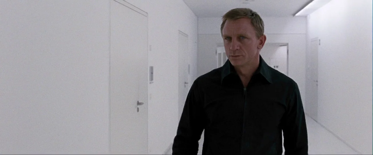 Quantum of Solace (2008) movie still: medium — A stark white, sterile corridor with multiple closed doors on both sides. A man wearing a black shirt…