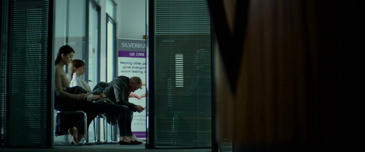 Wild Rose (2018) movie still: wide — A modern corridor features glass walls with aluminum frames and a promotional sign nearby. Three people sit on…