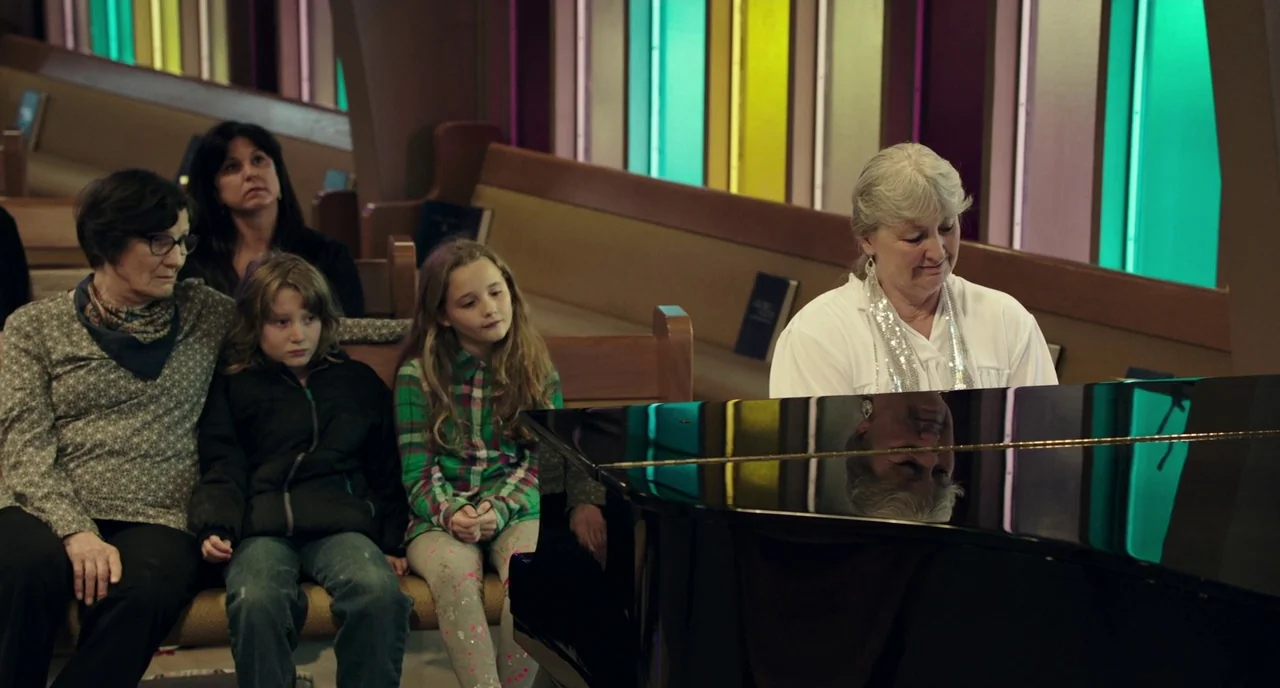 Leave No Trace (2018) movie still: medium — A row of wooden church pews is arranged before stained glass windows. A woman sits playing a grand piano…