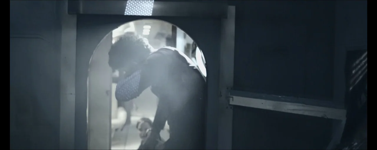 Turbulence (2025) movie still: medium — An interior shot shows a person standing within a curved doorway in a cabin. The individual leans forward…