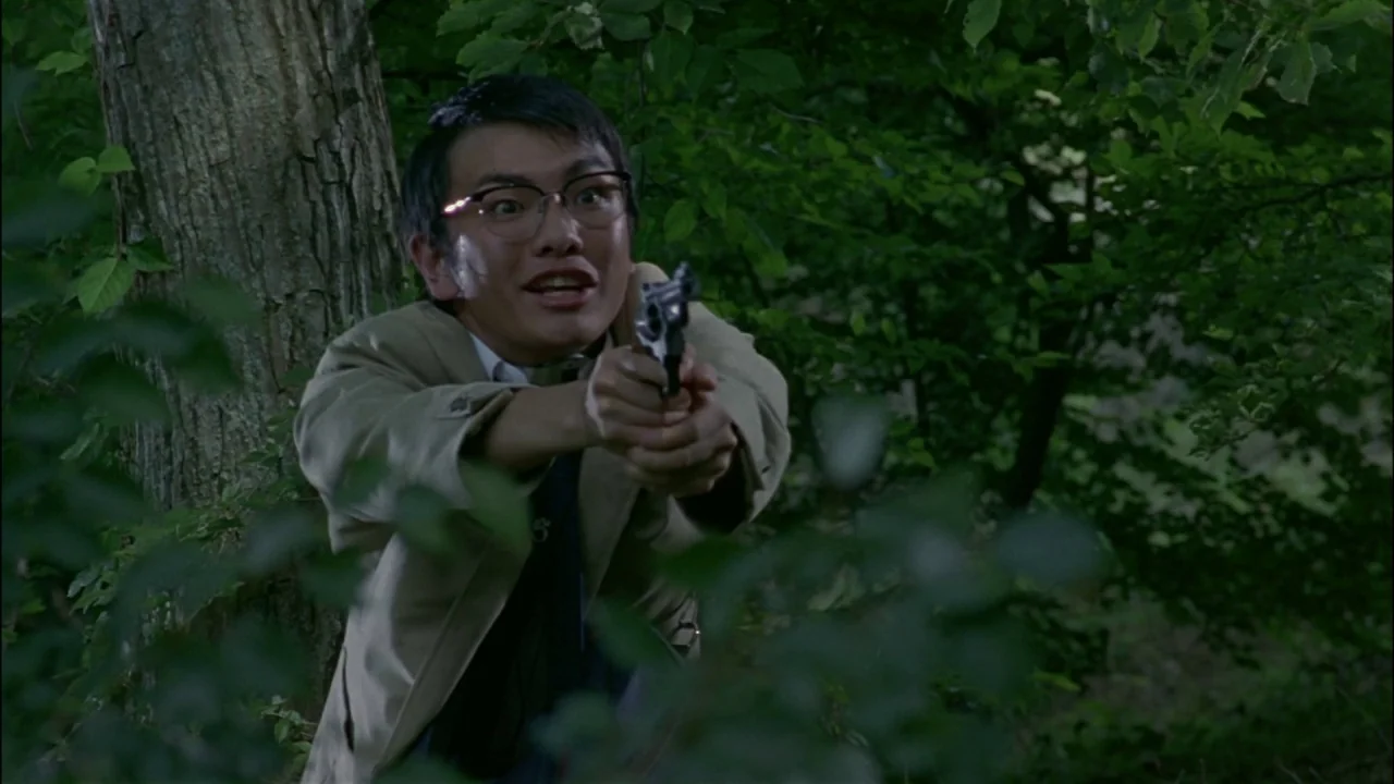 Battle Royale (2000) movie still: medium — A dense patch of woodland with thick foliage and a prominent large tree. A young man wearing a trench…