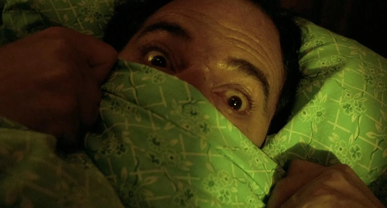 Delicatessen (1991) movie still: closeup — A man lies in bed with his face partially covered by a green floral sheet. He holds the patterned fabric…