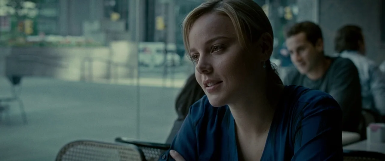 Limitless (2011) movie still: medium — A woman sits at a table near a large window looking out onto a street. She is positioned in the foreground…