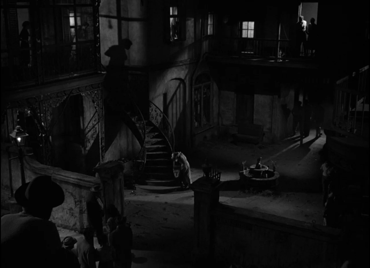 A Streetcar Named Desire (1951) movie still: wide — A dark, multi-level courtyard features a central stone fountain and a spiral iron staircase. A…