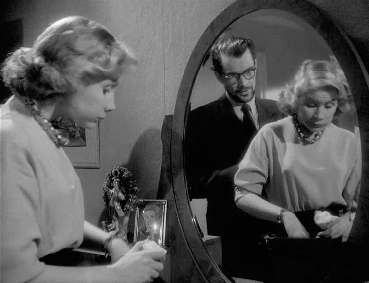 Prison (1949) movie still: medium — A woman stands before a vanity mirror while a man watches her from behind. The vanity top is cluttered with…