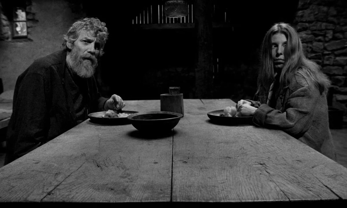 The Turin Horse (2011) movie still: medium — A rustic stone interior with heavy wooden walls and a large, worn dining table. A man and a woman sit…