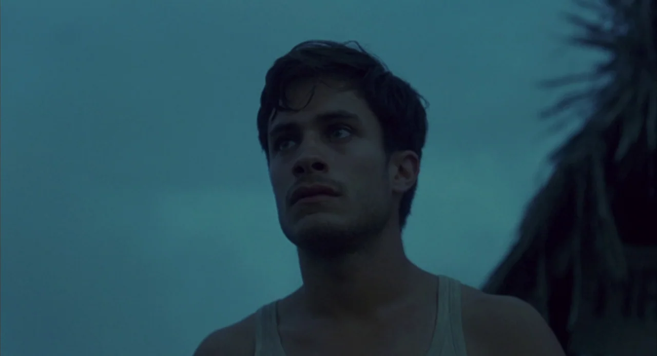 The Motorcycle Diaries (2004) movie still: medium — A man stands outdoors under a dark, overcast blue sky. The man looks toward the left of the…
