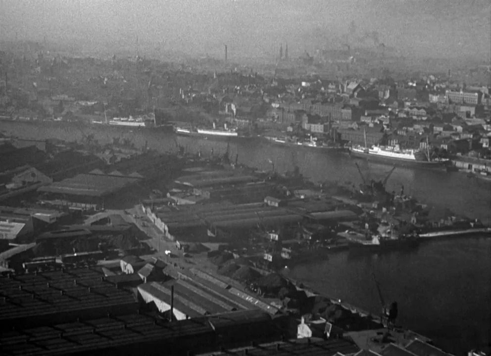 Odd Man Out (1947) movie still: establishing — An industrial harbor area features numerous low-profile warehouse buildings and docked cargo ships.…