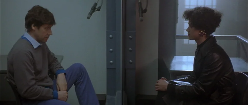 The Crying Game (1992) movie still: medium — A spartan visitation area is divided by a glass panel and metal frames. Two individuals sit on opposite…