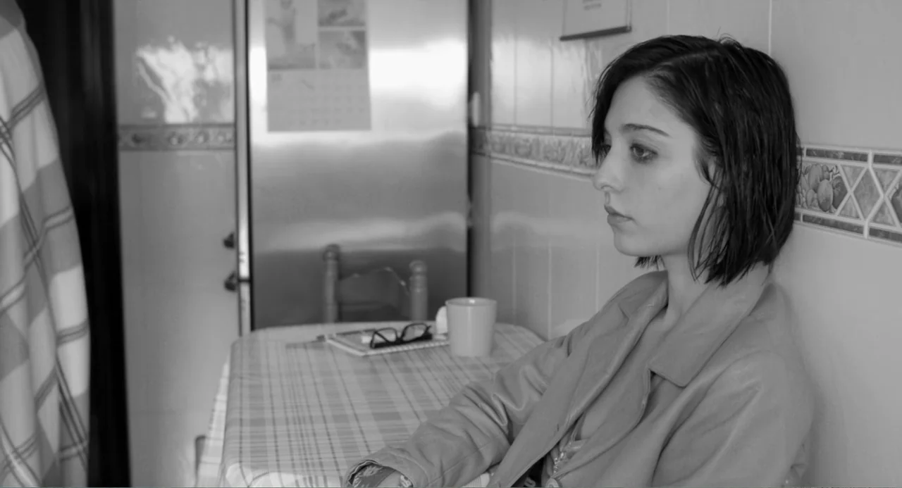 El Planeta (2021) movie still: medium — A modern kitchen interior features white tiled walls and a table covered with a checkered cloth. A young…