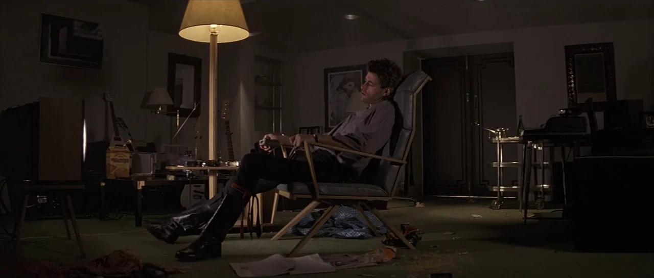 Pink Floyd The Wall (1982) movie still: wide — A sparsely furnished room with green carpet and various items scattered on the floor. A man rests in…