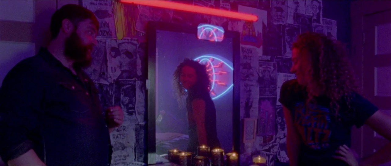 Bliss movie still: medium — A bedroom wall is densely layered with posters and illuminated by neon red lighting. A couple stands near a mirror…