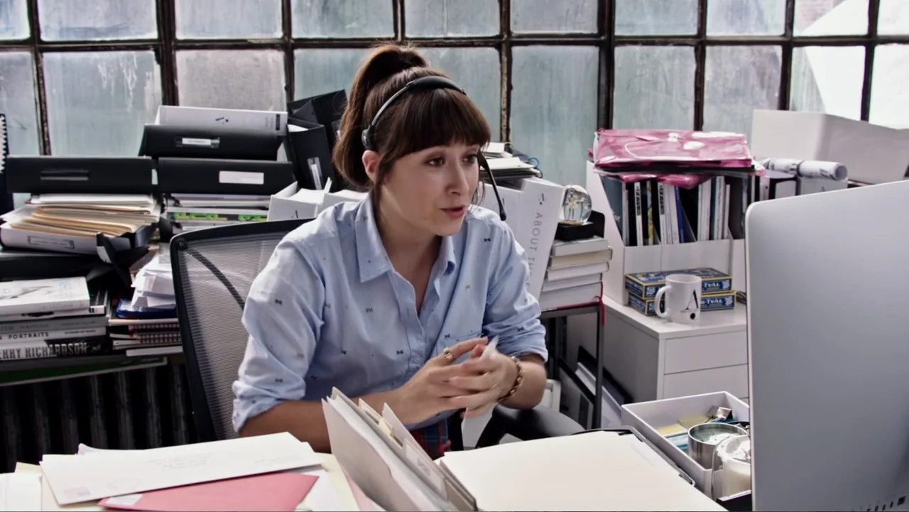 The Intern (2015) movie still: medium — A cluttered professional desk space is filled with piles of documents and folders. A young woman in a…